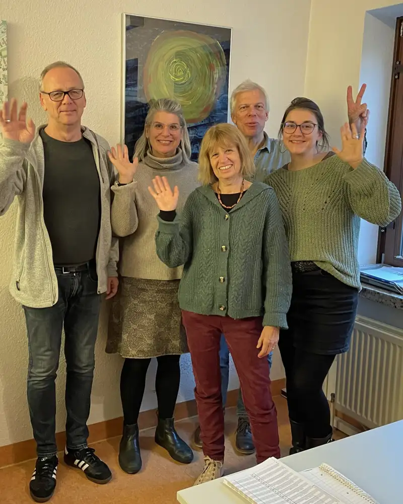 Gruppenbild mit drei Frauen und zwei Männern, die in einem Raum stehen und in die Kamera winken
