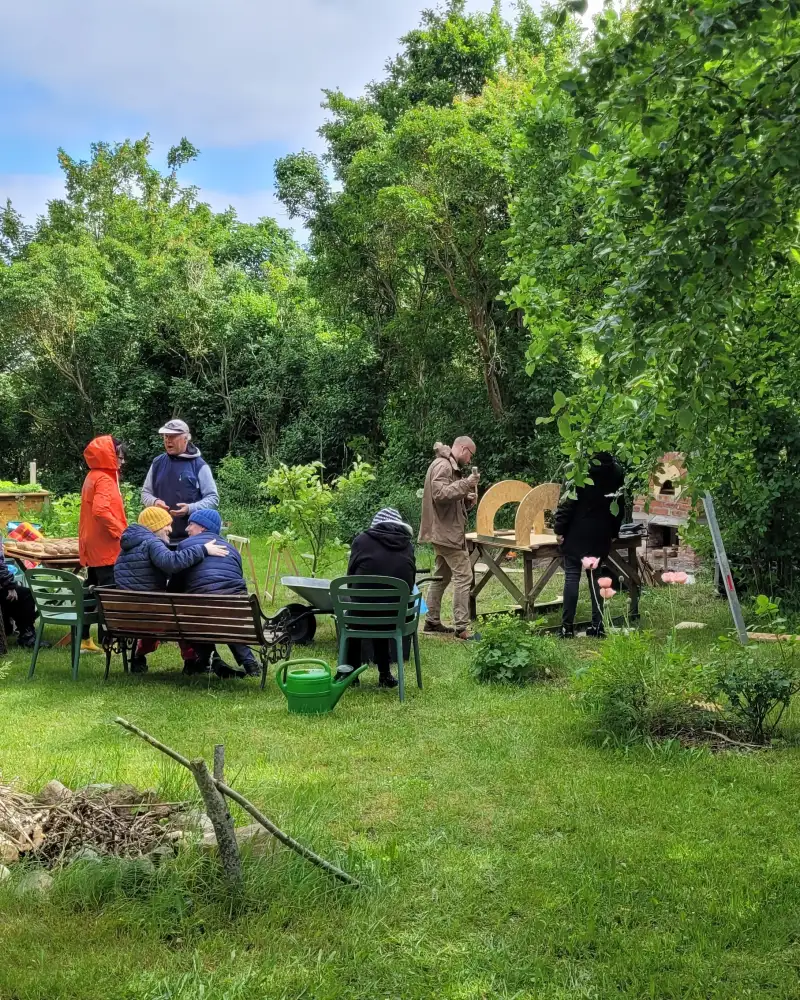 Eine Gruppe von neun Menschen sitzt im Halbkreis in einem Garten. Zwei von ihnen unterhalten sich stehend, zwei arbeiten beim Aufbau eines Lehmofens. Die meisten Personen sind von hinten zu sehen.