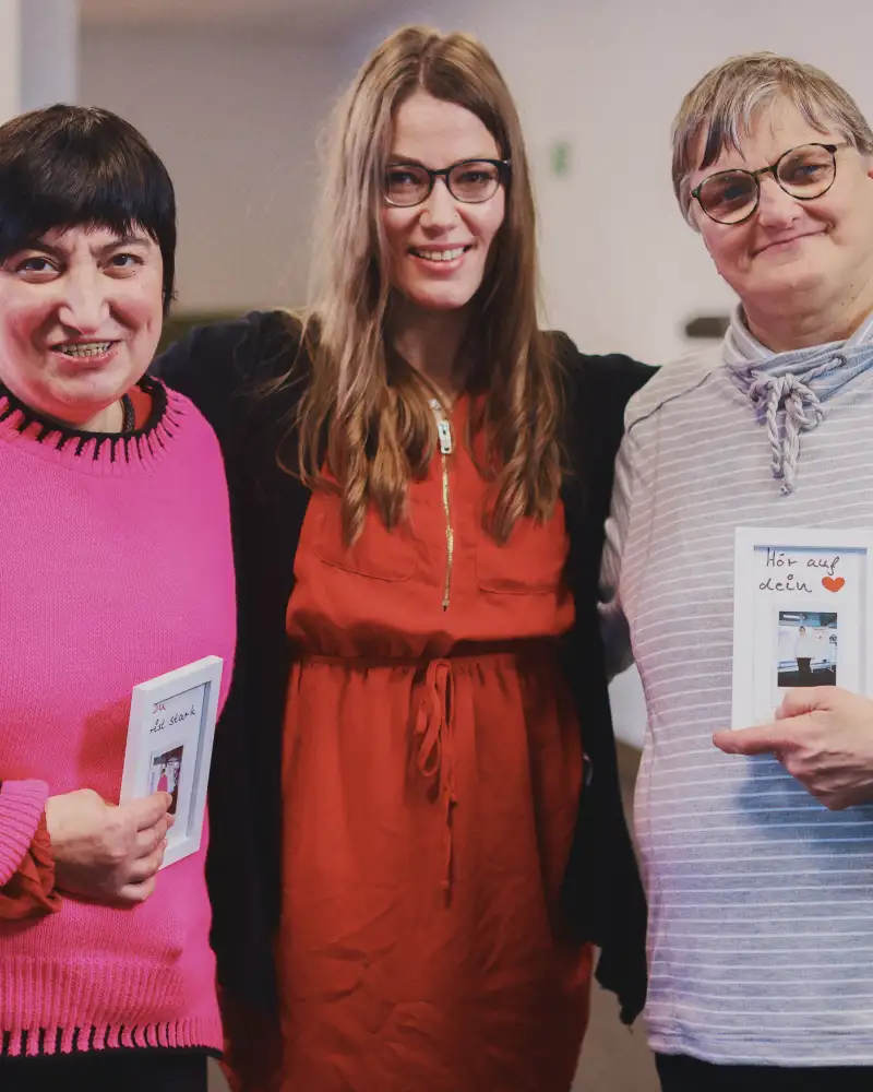 Drei Frauen stehen nebeneinander. Sie halten einen kleinen weißen Bilderrahmen in den Händen mit einem Foto von sich selbst und dem Satz "Hör auf dein Herz".