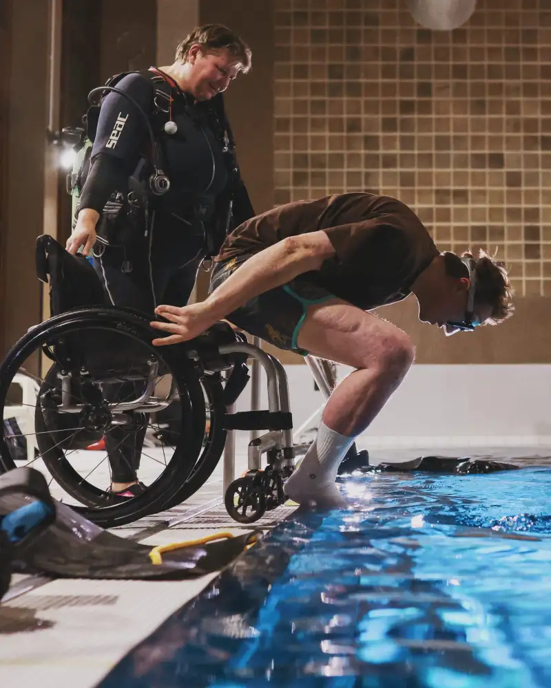 Ein Mann im Rollstuhl macht einen Köpper in ein Schwimmbecken. Eine Frau in einem Neoprenanzug hält den Rollstuhl fest. Ein Mann in Tauchausrüstung warten im Wasserbecken.