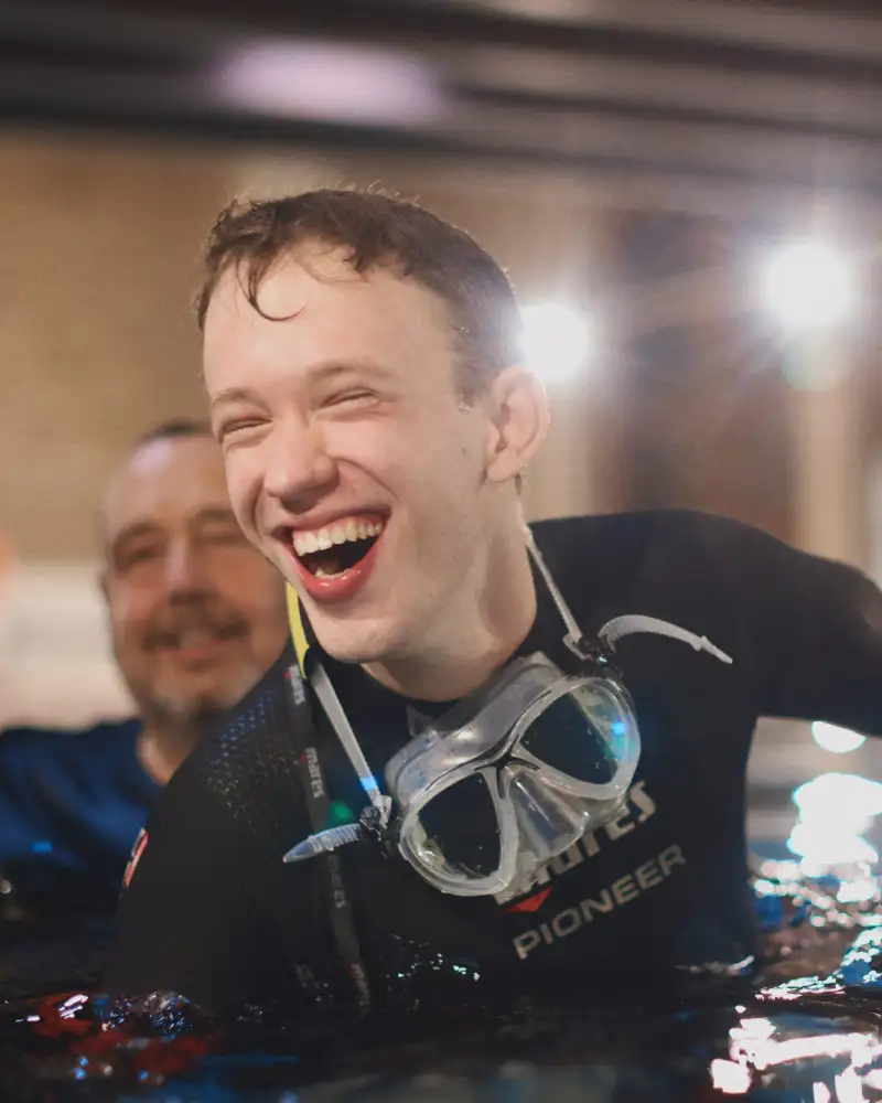 Drei Männer in Tauchkleidung schwimmen in einem Wasserbecken. Sie lachen.