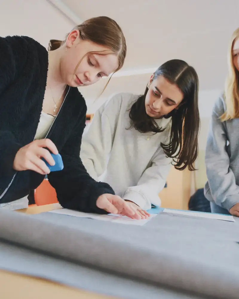 Drei junge Frauen zeichnen mit Nähkreide ein Muster auf ein Stück Stoff ein.