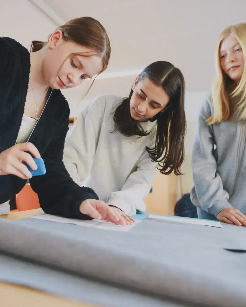 Drei junge Frauen zeichnen mit Nähkreide ein Muster auf ein Stück Stoff.