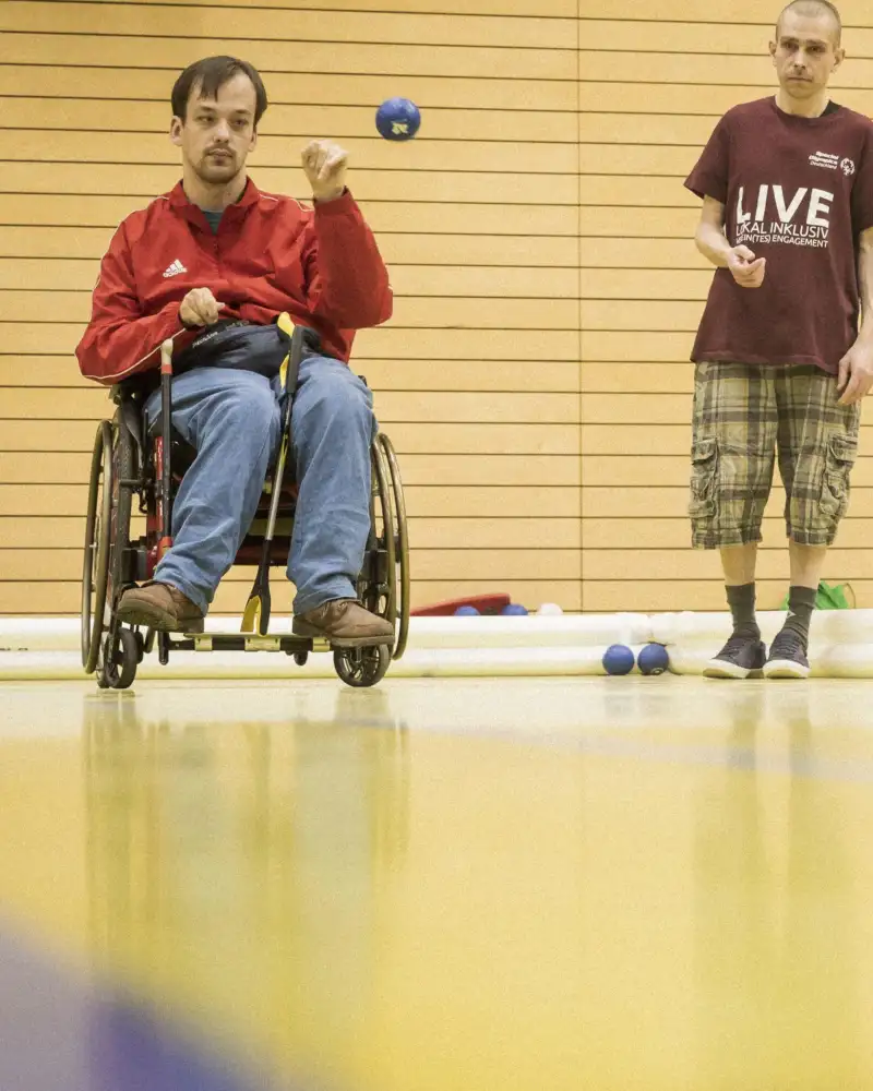 Ein Mann im Rollstuhl wirft eine Bocciakugel auf das Spielfeld.