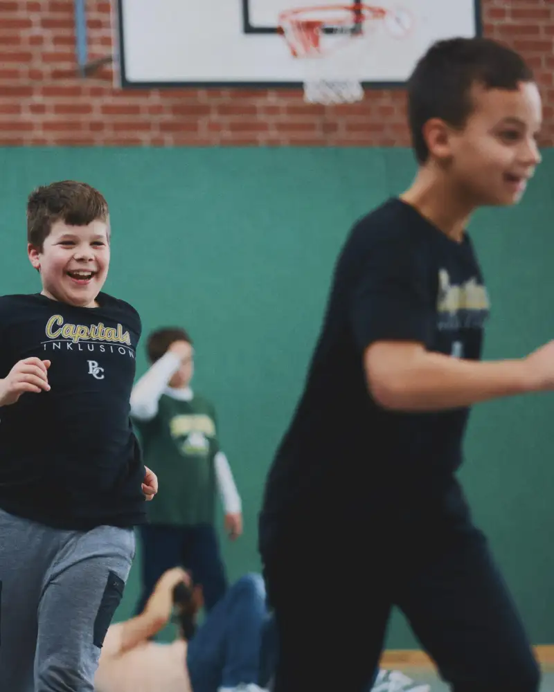 Zwei Jungen laufen lachend durch eine Sporthalle. Im Hintergrund sind weitere Kinder zu sehen.