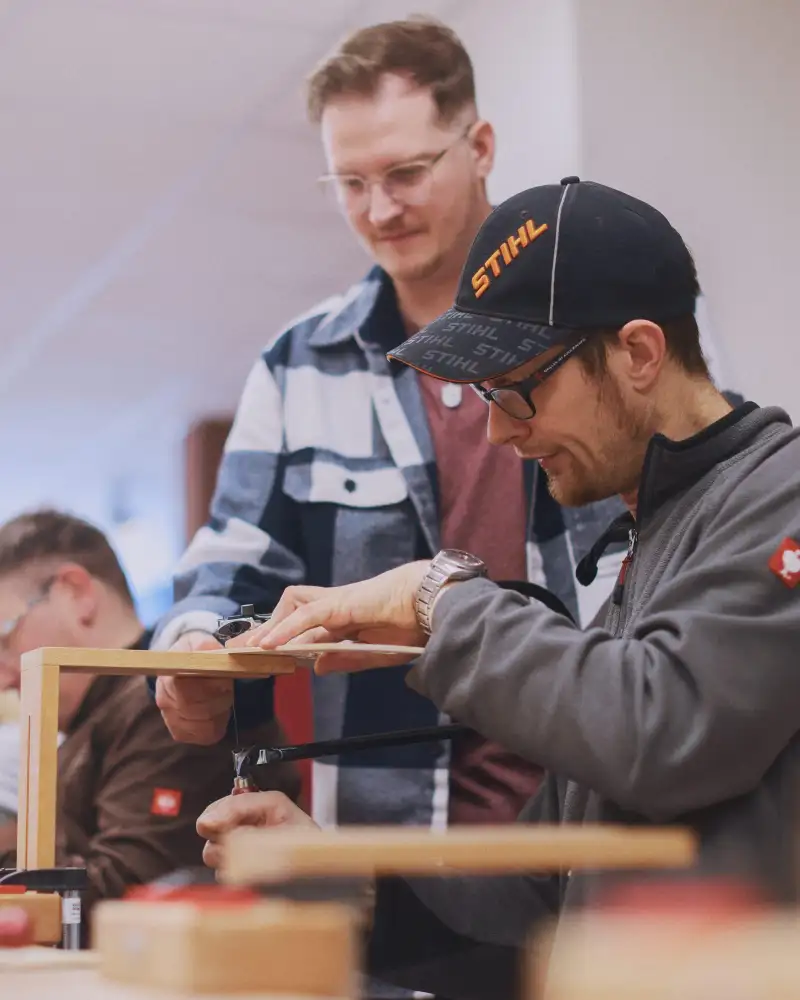 Ein junger Mann mit Kappe sägt eine Schablone aus. Ein anderer hält das Werkstück fest.