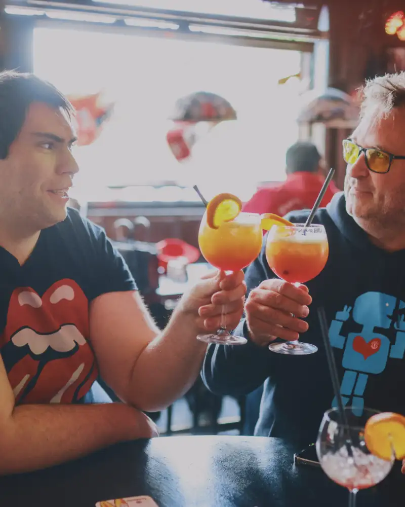 Zwei Männer an einem Stehtisch stoßen mit orangefarbenen Cocktails an.