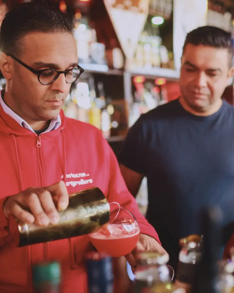 Ein Mann mit Brille schüttet einen Cocktail aus dem Shaker in ein Glas. Er steht hinter einer Theke. Neben ihm steht ein weiterer Mann.