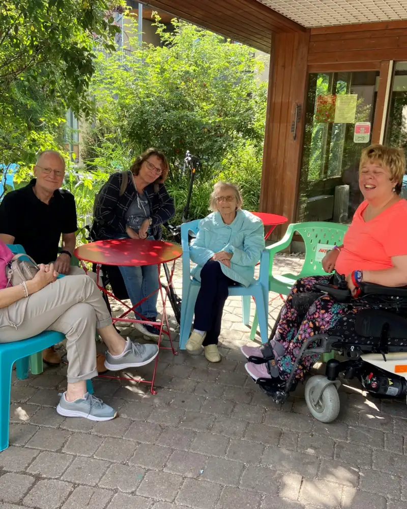 Fünf Menschen mit und ohne Behinderung sitzen im Halbkreis auf einer Veranda