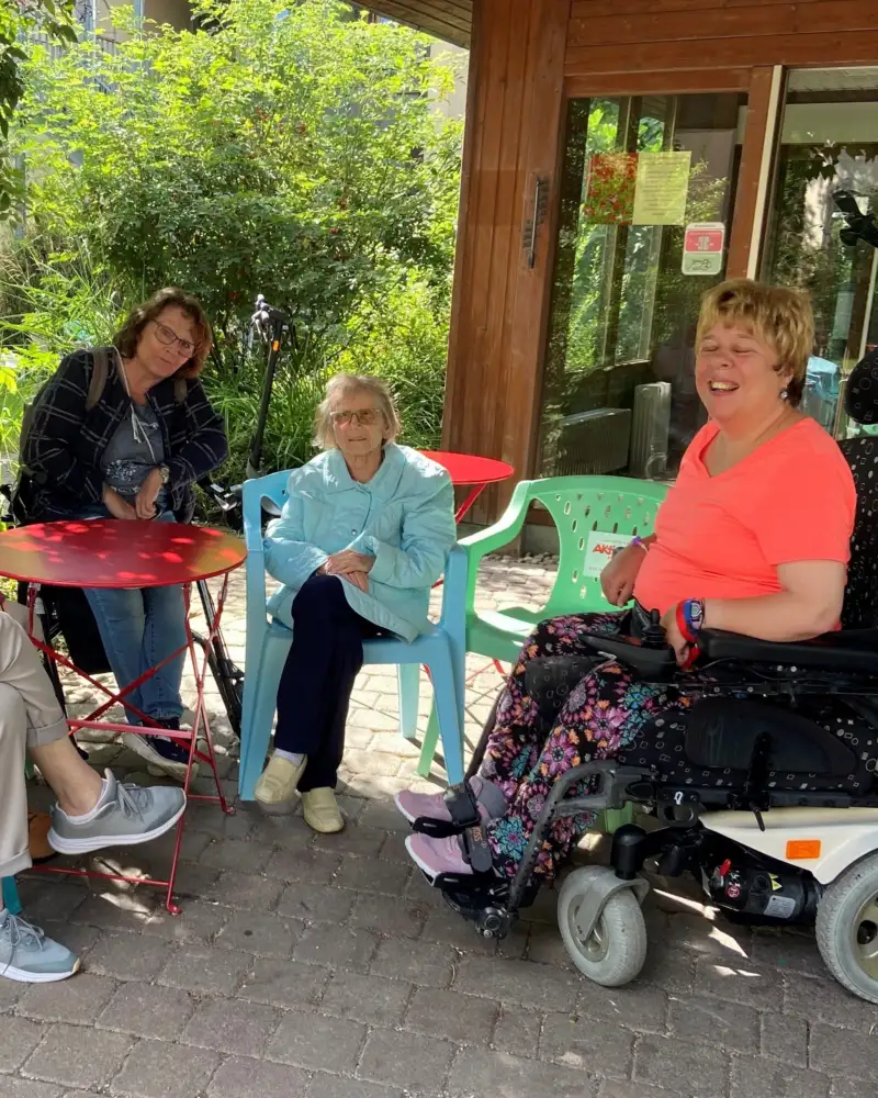Eine Gruppe von Personen mit und ohne Behinderung sitzen im Halbkreis auf einer Terrasse.