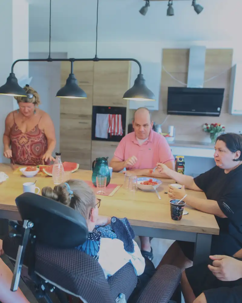 Sieben Menschen mit und ohne Behinderung sitzen um einen großen Küchentisch und essen Wassermelone.