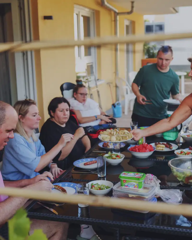 Sieben Menschen mit und ohne Behinderung sitzen an einem großen Tisch auf einer Terrasse und essen Salat.