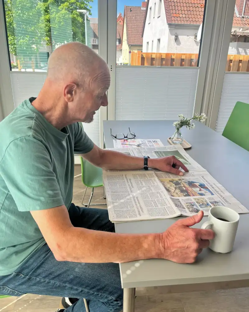 Ein Mann sitzt mit einer Tasse am Tisch und liest in einer Zeitung.