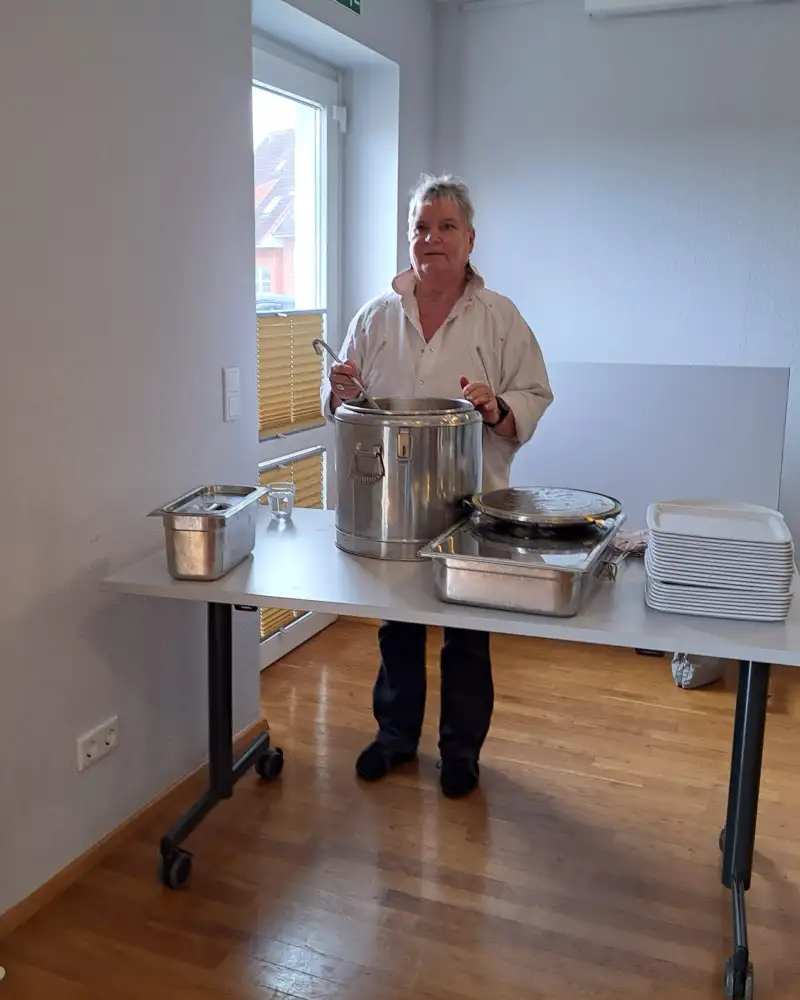 Eine Frau mittleren Alters steht in einem Raum hinter einem Tisch, auf dem ein sehr großer Kochtopf, verschiedene Metallbehälter und ein Stapel Tabletts stehen. Sie hält eine Schöpfkelle in der Hand.