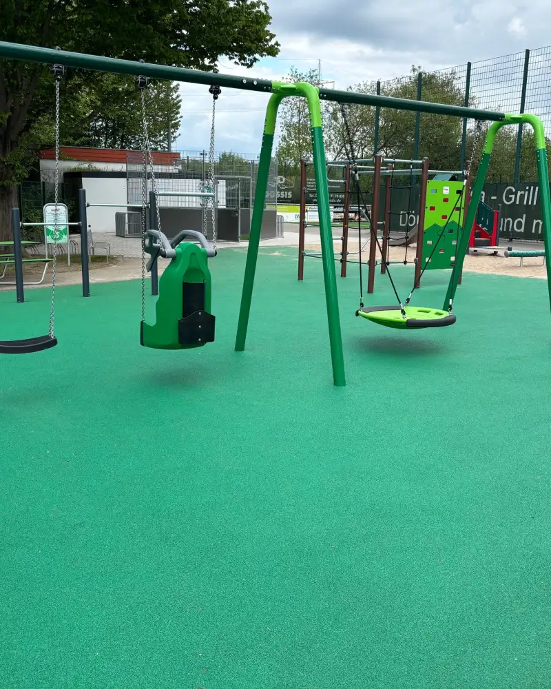 Auf einem Spielplatz, der mit einem grünen Belag ausgelegt ist, steht ein Gerüst. Daran hängen drei unterschiedliche Schaukeln. Daneben steht ein weiteres Gerät, das aussieht wie eine große bewegliche Schale mit einer hohen Kuppel, die aus drei Streben besteht.
