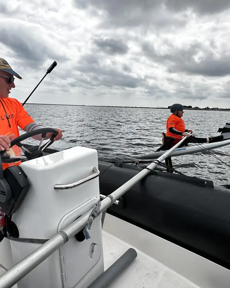 Zwei Männer in orangefarbenen Warnwesten sitzen hintereinander auf einem Motorboot. Der vordere Mann steuert das Boot. Am Boot ist eine lange Stange angebracht, die auf die See hinausragt. An ihrem Ende ist ein Surfbrett befestigt, das auf einem Tragflügel aus dem Wasser emporragt. Darauf sitzt ein Mann. Er ist gesichert und trägt ebenfalls eine Warnweste.