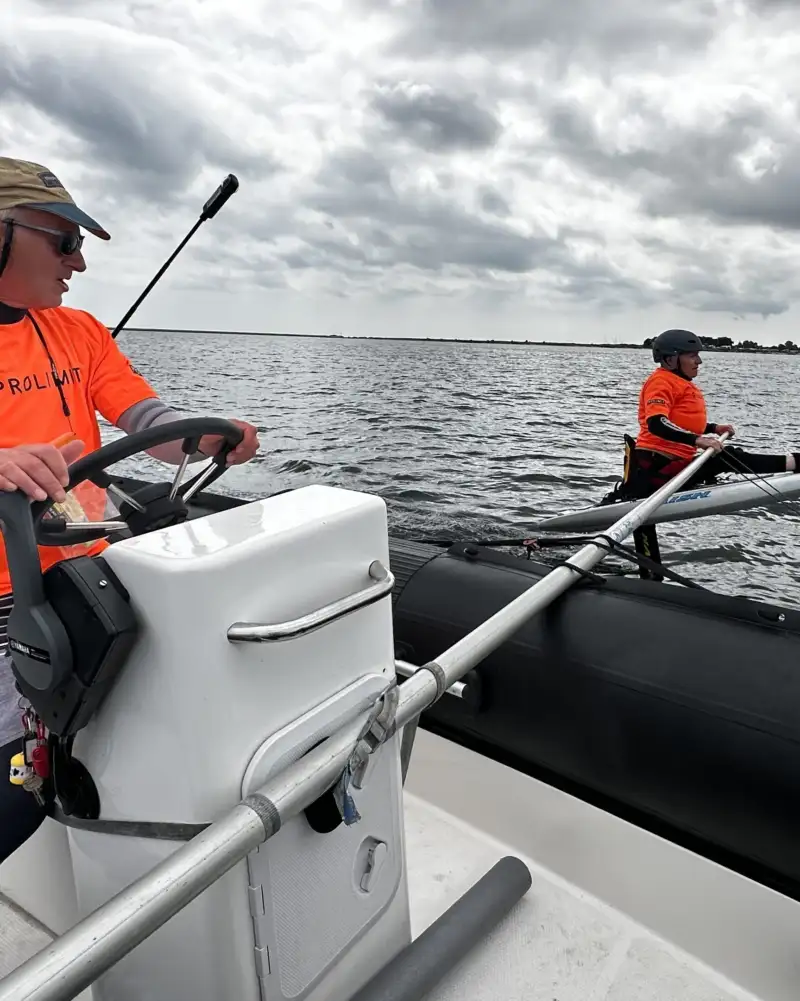 Zwei Männer in orangefarbenen Warnwesten sitzen hintereinander auf einem Motorboot. Der vordere Mann steuert das Boot. Am Boot ist eine lange Stange angebracht, die auf die See hinausragt. An ihrem Ende ist ein Surfbrett befestigt, das auf einem Tragflügel aus dem Wasser emporragt. Darauf sitzt ein Mann. Er ist gesichert und trägt ebenfalls eine Warnweste.