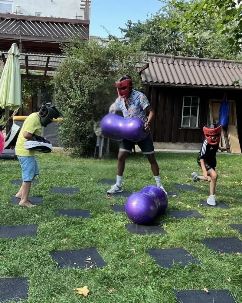 Drei Kinder spielen auf einer Wiese mit Bällen. Im Hintergrund stehen niedrige Gebäude. Die Kinder tragen Masken. Eines der Kinder hat eine dunkle Hautfarbe.