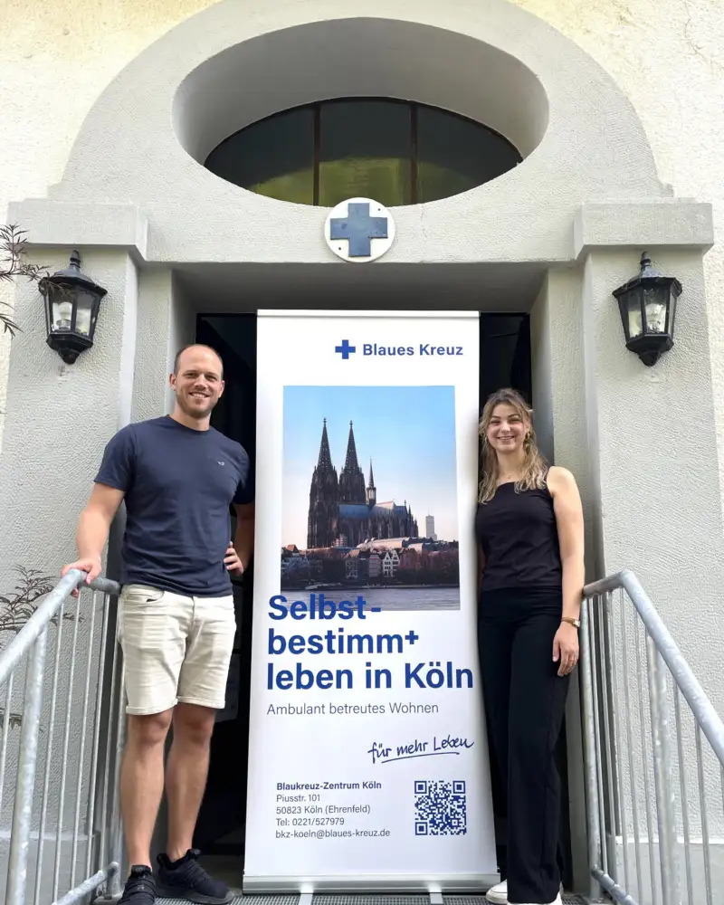 Ein Mann und eine Frau stehen in einem Hauseingang mit einem Plakat, auf dem u.a. steht "Selbstbestimm+ leben in Köln"