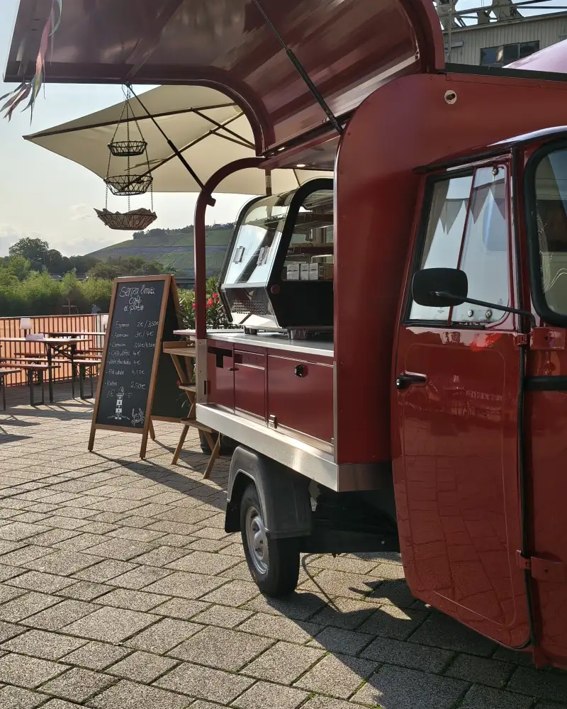 Ein Fahrzeug mit einer Kaffeebar parkt auf einer Terrasse. Im Hintergrund stehen Tische und Bänke.
