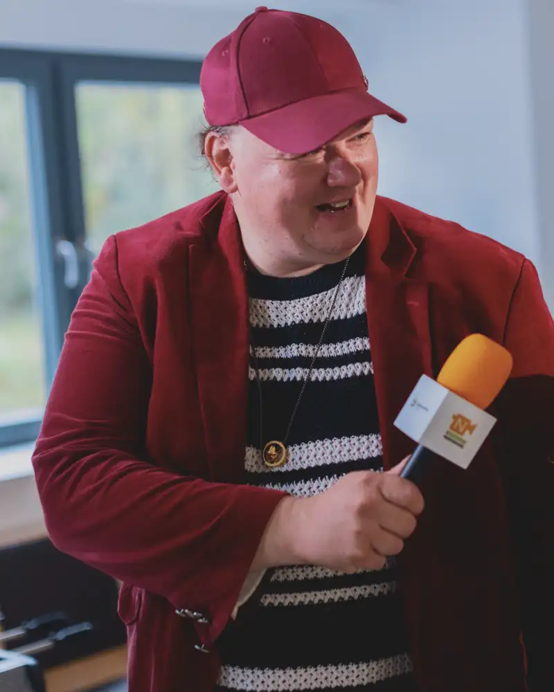 Ein großer Mann mit Sakko, grob gestrickten schwarz-weiß-gestreiftem Pulli und einem Cappy hält einer jungen Frau mit langen, gelockten roten Haaren und hellem geripptem T-Shirt ein Mikrofon hin, in das sie hineinspricht. Die beiden stehen in einem etwas kahlen Raum mit weißen Wänden.