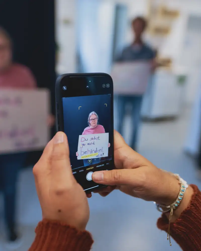 Ein Mensch trägt ein beschrifteten Zettel in der Hand und wird dabei mit einem Handy gefilmt