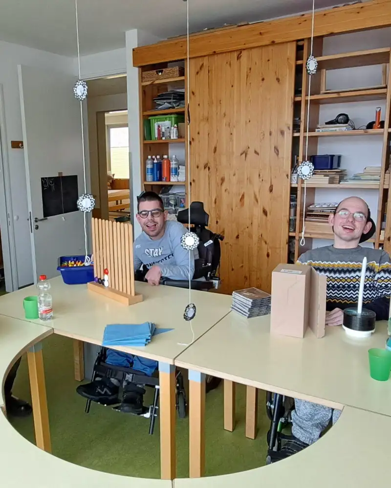 Zwei Männer sitzen im Rollstuhl an einem Tisch, auf dem verschiedene Materialien stehen