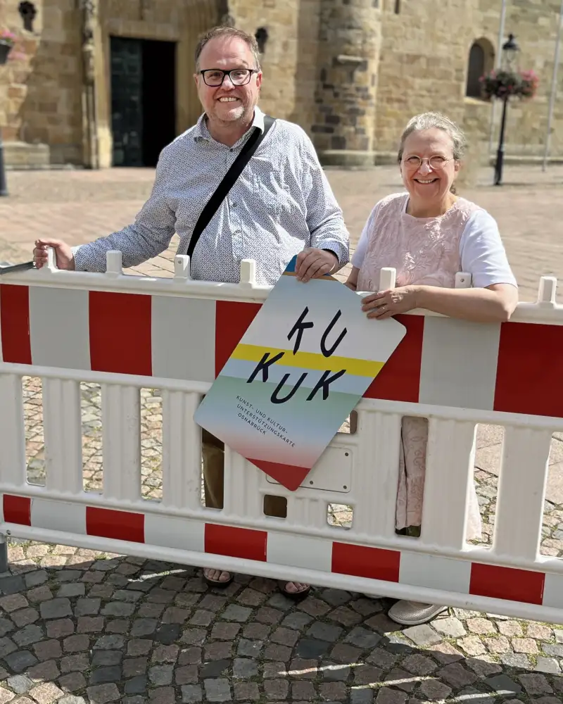 Ein Mann und eine Frau stehen hinter einer Absperrung auf einem Platz. Sie tragen ein Schild mit der Aufschrift "KUKUK".