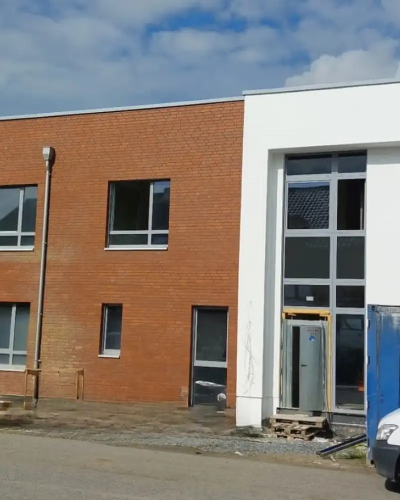 An einer Straße stehen vier Reihenhäuser mit Flachdächern, von denen nur die mittleren beiden vollständig auf dem Bild sind. Ihre Fassaden sind im Wechsel weiß gestrichen und mit roten Klinkern versehen. Die weißen Häuser stehen ein wenig vor. Auf der Straße stehen zwei weiße Lieferwagen.