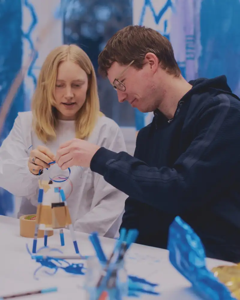 In einem Atelier basteln ein junger Mann im Hoodie und eine junge Frau in weißem Kittel und blonden Haaren zusammen ein Ding, dass wie eine Rakete aussieht. Auf dem Tisch stehen viele Bastel-Utensielien. Die Wände im Hintergrund sind blau bemalt.