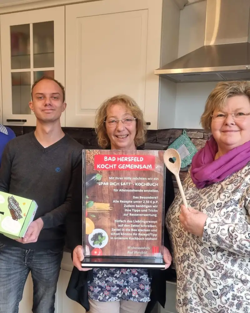Drei Frauen und ein Mann stehen nebeneinander in einer Küche und lächeln in die Kamera. Eine von ihnen hält einen Kochlöffel  hoch, eine andere trägt einen Aufsteller mit einem Plakat, darauf steht: Bad Hersfeld kocht gemeinsam. Die dritte Frau hält ein weißes Din-A-4-Blatt hoch. Der Mann hat einen länglichen Karton in den Händen.