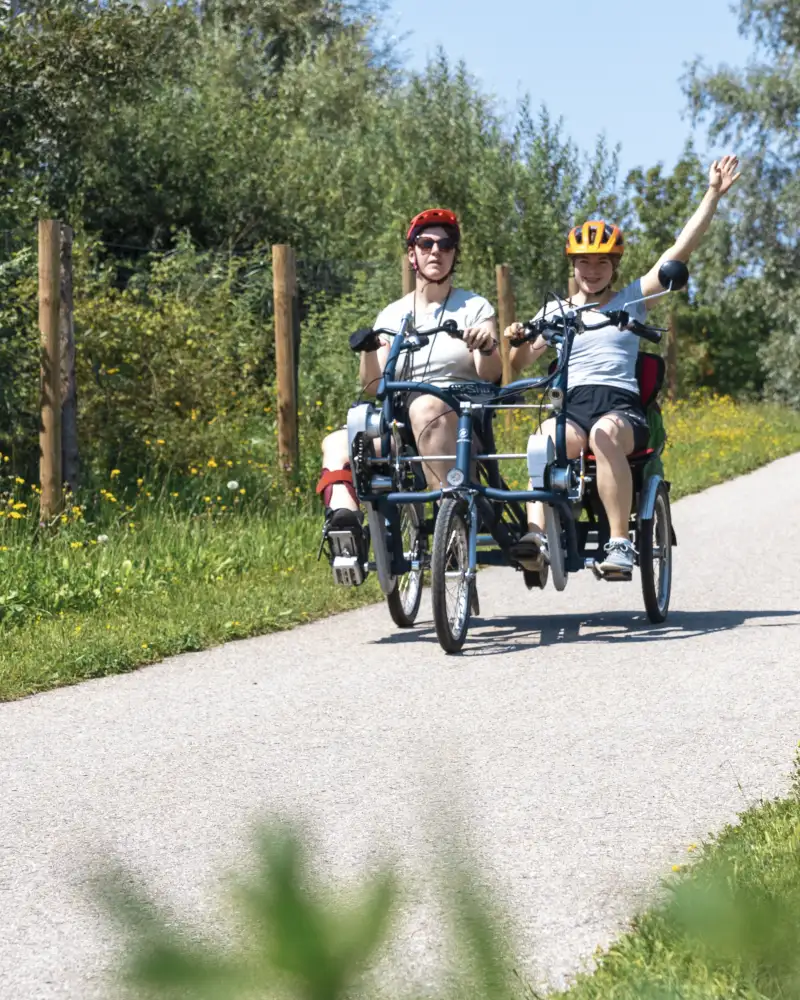 Zwei Personen sind auf einem gut ausgebauten Waldweg mit einem Elektro-Tandem unterwegs. Sie sitzen nebeneinander und tragen Fahrradhelme. Hinter ihnen nähert sich ein weiteres Gefährt.
