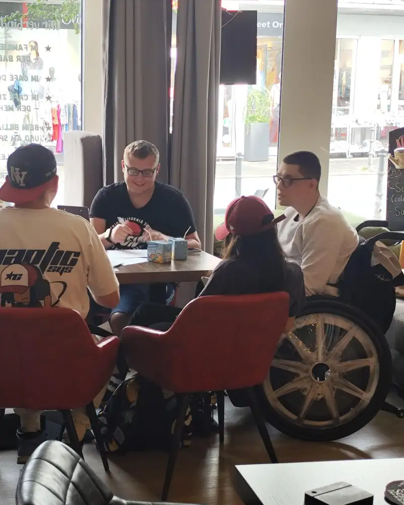 Eine junge Frau und drei junge Männer sitzen an einem Tisch vor großen Fenstern mit Blick auf eine Straße. Einer der jungen Männer sitzt im Rollstuhl.