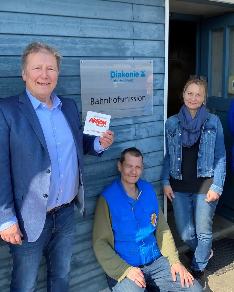 Eine Frau und zwei Männer neben dem Eingang zu einer Bahnhofsmission. Einer der Männer hält eine Karte mit der Aufschrift "Aktion Mensch" hoch. Der zweite Mann sitzt auf dem Boden.