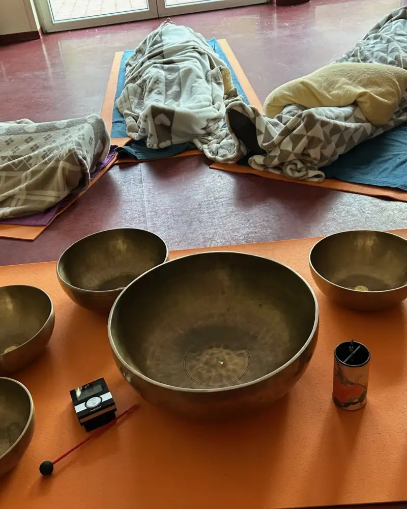 In einem Raum liegen mehrere Personen in Wolldecken gehüllt auf Matten. Vor ihnen steht ein sehr flacher Tisch, auf dem Klangschalen in verschiedenen Größen platziert sind.
