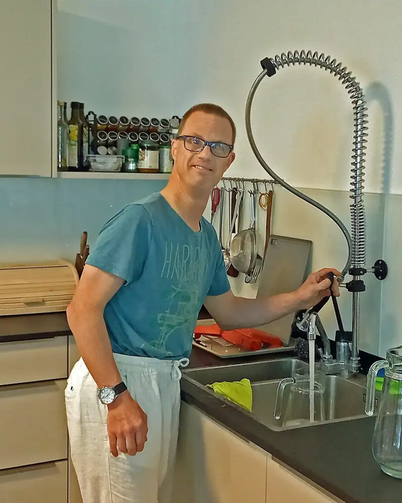 Ein Mann steht an der Spüle einer Küche. Er trägt ein T-Shirt und eine Brille.