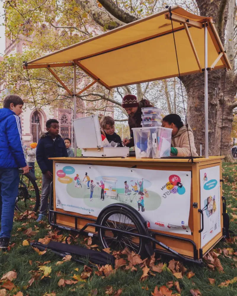 Vier Kinder und ein Mann stehen um einen mobilen Stand, auf dem in transparenten Kisten Malutensilien gestapelt sind.