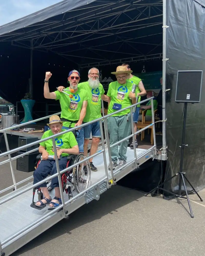 Fünf Männer in grünen T-Shirts stehen auf einer Rampe vor einer Bühne, einer davon sitzt im Rollstuhl