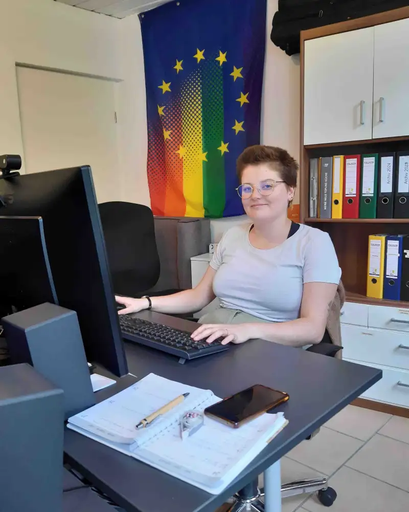 Eine junge Frau sitzt an einem Schreibtisch vor einem Monitor. Hinter ihr an der Wand hängt eine Fahne in Regenbogenfarben.