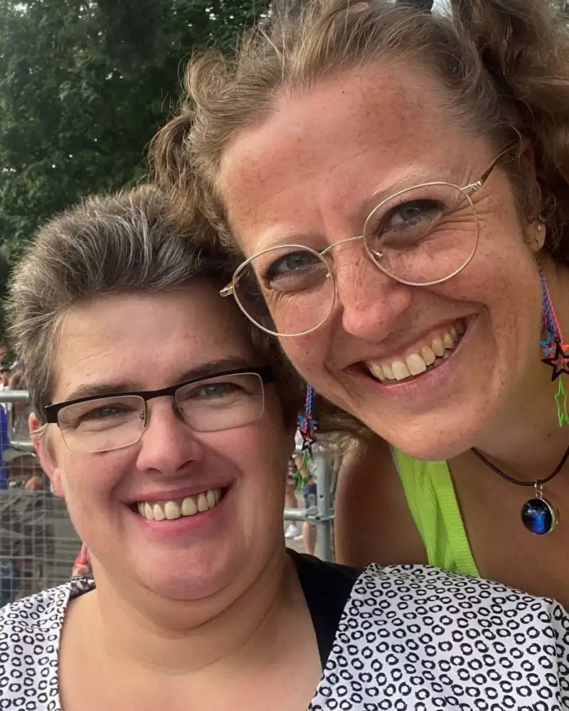 Zwei Frauen schauen lächelnd in die Kamera. Im Hintergrund stehen Menschen hinter einer Absperrung.