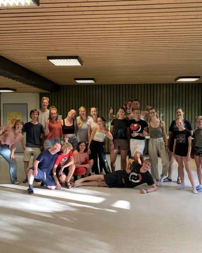 Eine Gruppe von 20 Jungen und Mädchen stehen in einem großen Raum mit Holzgetäfelter Decke. Einer von ihnen hat das Down-Syndrom.