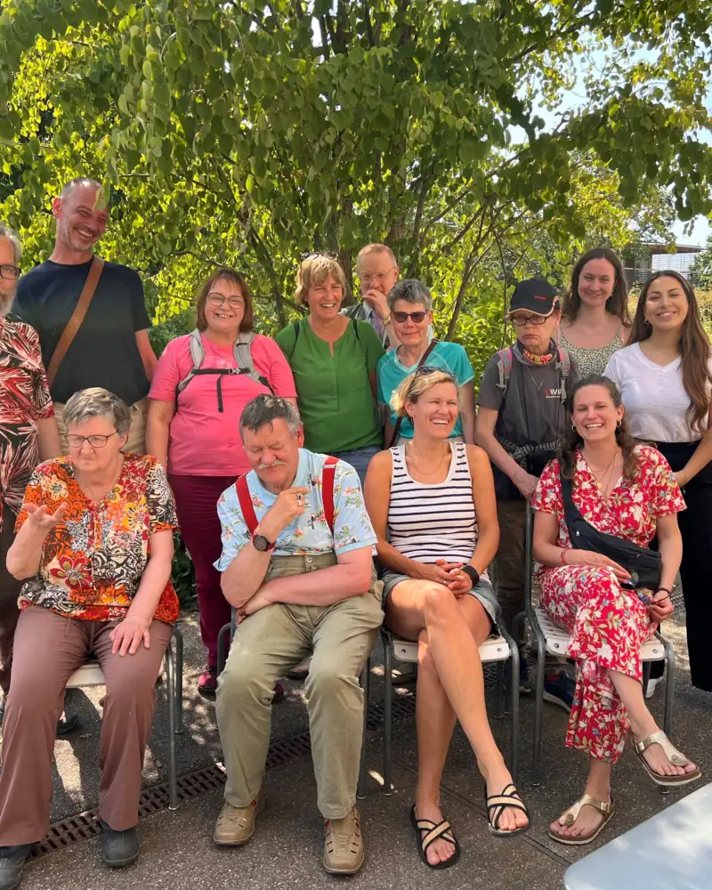 Vier Männer, ein Junge und acht Frauen stehen und sitzen im Schatten eines Baumes und lächeln in die Kamera.