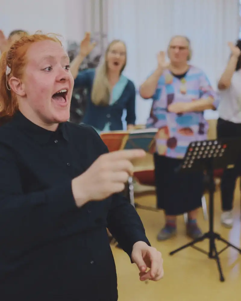 Gebärdenchor bei der Probe. Vorne steht eine junge Frau, die den Takt angibt, im Hintergrund stehen sechs Persondn in einer Reihe. Sie haben Notenpulte vor sich und heben ihre rechte Hand.