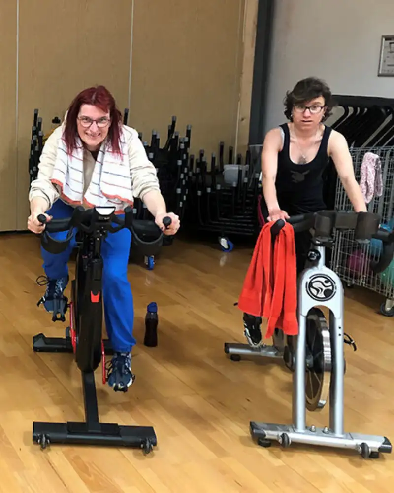 Mehrere Frauen mit und ohne Behinderung sitzen nebeneinander auf Heimtrainern.