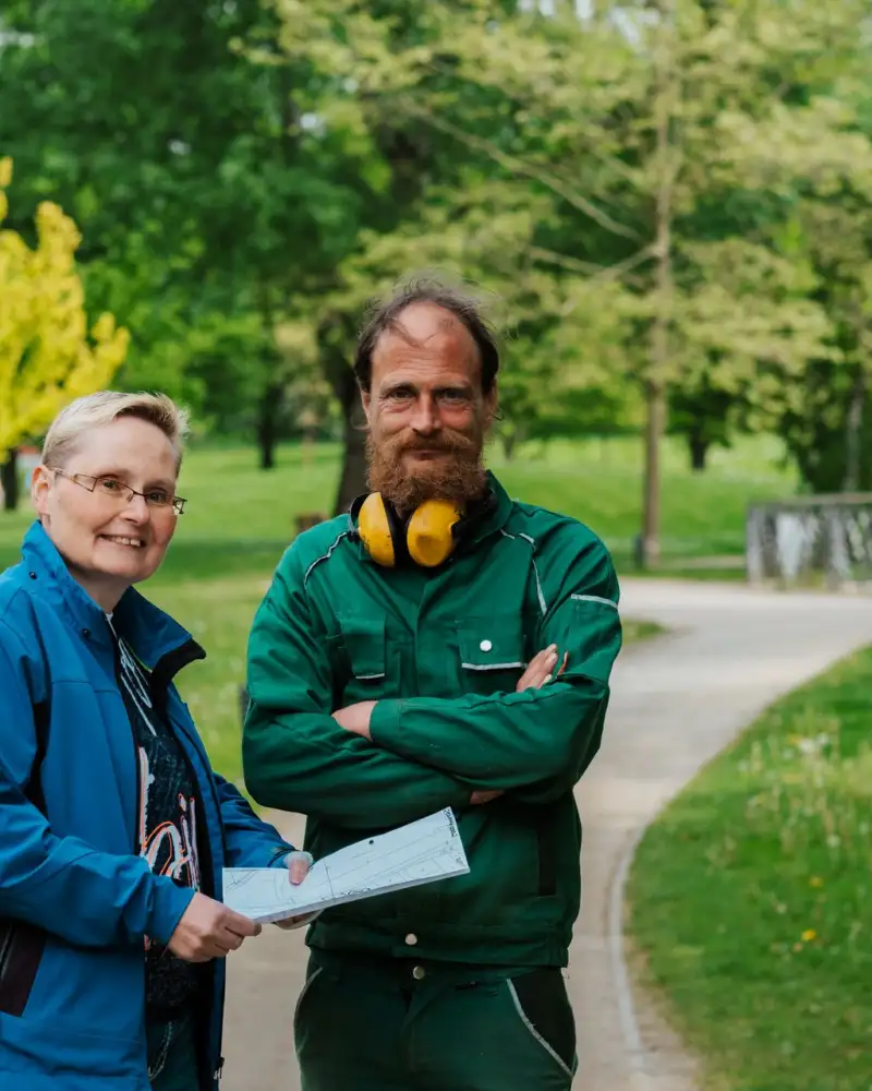 Ein Mann und eine Frau stehen in einem Park. Der Mann trägt einen grünen Overall. Er hat Kopfhörer.