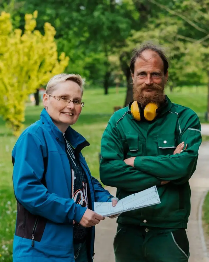 Ein Mann und eine Frau stehen in einem Park. Der Mann trägt einen grünen Overall und Kopfhörer um den Hals.