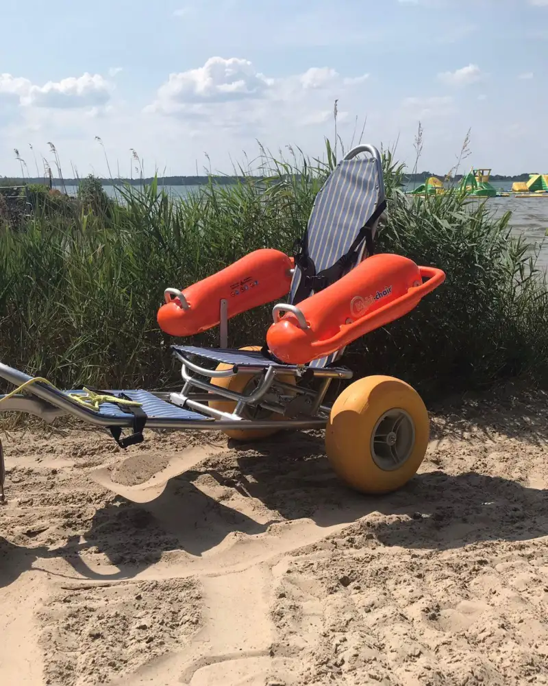 Ein Rollstuhl auf drei kleinen dicken Rädern (zwei hinten, eines vorne) steht auf einem Sandstrand, im Hintergrund die See.