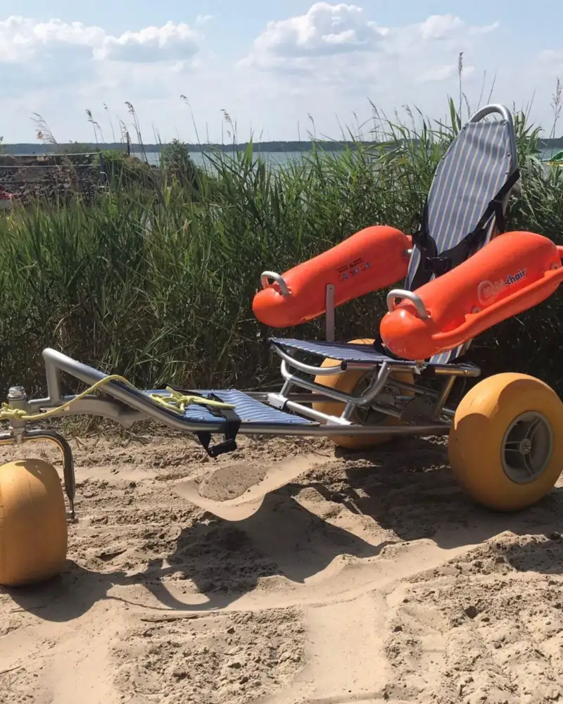 Ein Rollstuhl an einem Sandstrand, der auf drei kleinen dicken Rädern (zwei hinten, eines vorne) steht. Im Hintergrund die See.