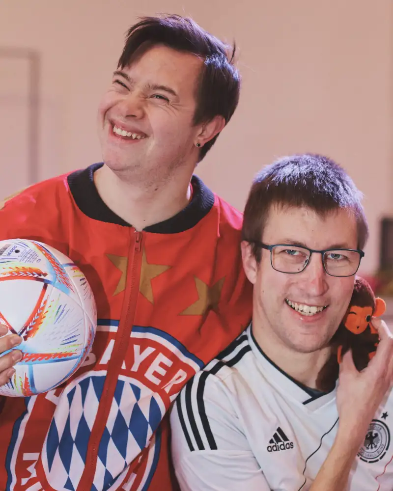 Zwei junge Männer, einer mit Downsnyndrom, mit Fußball-T-Shirt und einem Ball in einem Raum.