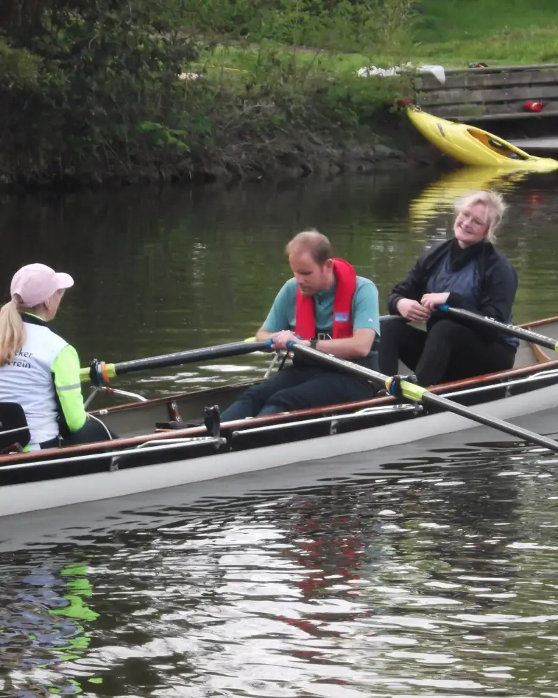 Drei Personen in einem Ruderboot auf einem Gewässer.